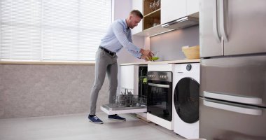 Happy Man Arranging Plates In Dishwashing Machine In Kitchen