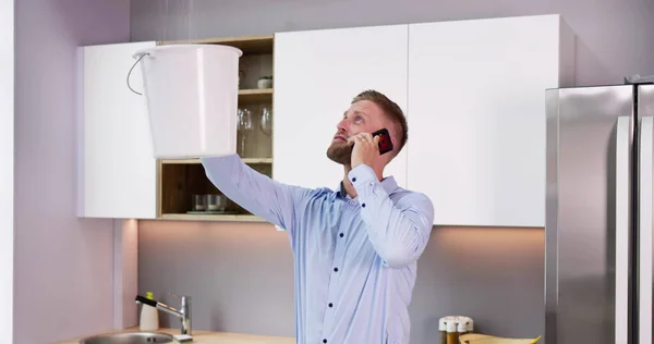 Man Calling Plumber While Collecting Water Leaking Ceiling Using Bucket — Stock Photo, Image