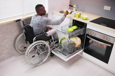 Tekerlekli sandalyede oturan ve bulaşık makinesindeki tabakları düzenleyen genç adam.