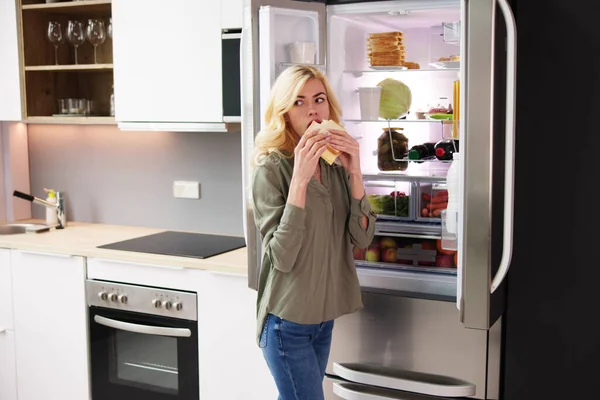 オープン冷蔵庫の近くでチーズサンドを食べる空腹の女性 — ストック写真