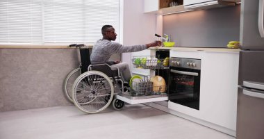 Tekerlekli sandalyede oturan ve bulaşık makinesindeki tabakları düzenleyen genç adam.