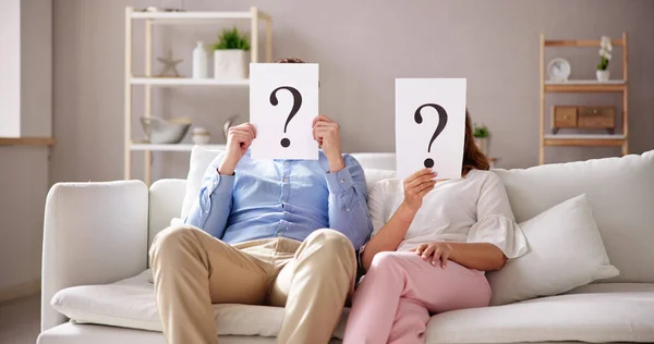 Couple Sitting On Sofa Holding Question Mark Sign In Front Of Face