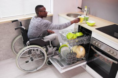 Tekerlekli sandalyede oturan ve bulaşık makinesindeki tabakları düzenleyen genç adam.