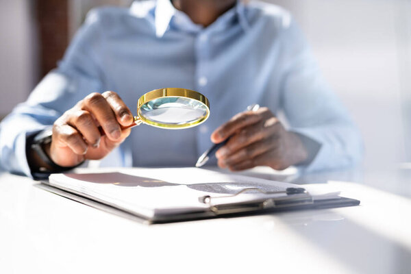 Close-up Of A Businessperson's Hand Holding Magnifying Glass Over Invoice At Workplace