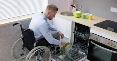 Mutfakta bulaşık makinesi kullanan tekerlekli sandalyedeki engelli kişi.