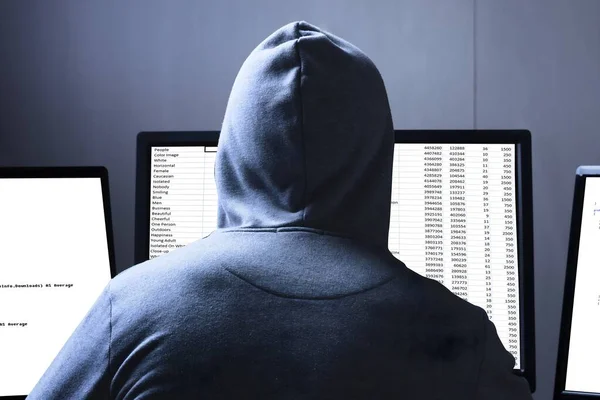 Rear View Of A Hacker Using Multiple Computers For Stealing Data On Desk