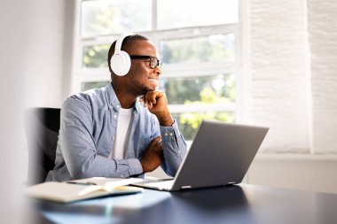 Genç Afrikalı Amerikalı öğrenci online olarak dizüstü bilgisayar ve kulaklıkla çalışıyor, eğitimde giyim kuşaklarının keyfini çıkarıyor ve sınavlara hazırlanıyor. Muhtemel serbest öğretmen..