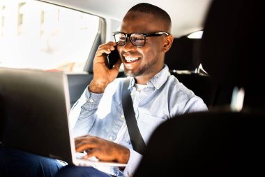 Afrikalı işadamı lüks bir arabada oturmuş bilgisayarında Amerikan yöneticisiyle online iş maceralarını tartışırken akıllı telefondan konuşuyor..
