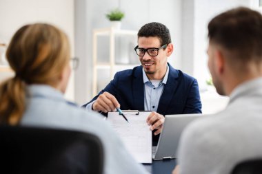 Finansal danışman ofisteki genç çifte yeni proje yatırımını açıklıyor. Emlakçı aileyle ipotek seçeneklerini tartışıyor. Finans danışmanıyla ipotek kredisi danışmanlığı.