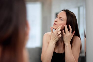 Endişeli dermatolog aynada genç bir kadının yüzündeki sivilceleri inceliyor..