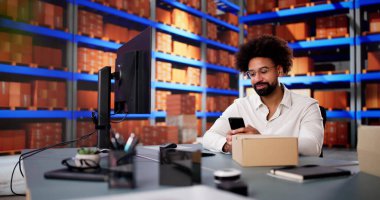 Afrikalı Amerikalı adam küçük bir ofis deposunda akıllı telefonuyla barkodları tarıyor..