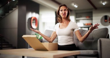 Kadın oturma odasında kırık paket kutusunu üzgün bir ifadeyle açıyor. Muhtemel Meslekler: Teslimat Kişi