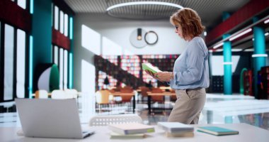 Üniversite kütüphanesinde dizüstü bilgisayar ve kitapla araştırma yapan kendini adamış kadın profesör..