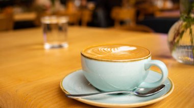 Açık mavi seramik bir fincanda Rosetta tarzı süt köpüğüyle bir fincan latte ve sıcak aydınlatılmış bir kafe içinde ahşap bir masa üzerinde çay tabağı..