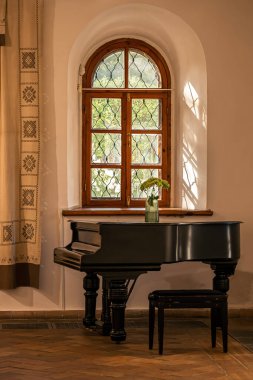 Grand piano with bench placed under arched wooden window and traditional curtain, interior detail with flowers and sunlight, cultural and musical heritage concept.