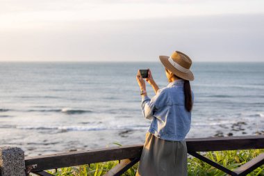 Gezgin kadın gün batımında deniz kenarında fotoğraf çekmek için cep telefonu kullanıyor.