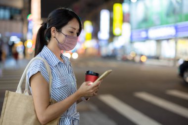 Yüz maskeli ve cep telefonu kullanan ve geceleri sokakta yürüyen bir kadın.