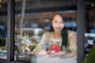 Kadın cep telefonu kullanıyor ve kahve dükkanında cam yansıması ile oturuyor.