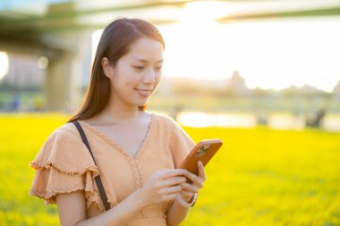 Kadın parkta güneş ışığı altında cep telefonu kullanıyor. 