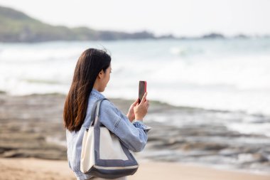 Kadın deniz kenarında fotoğraf çekmek için cep telefonu kullanıyor.