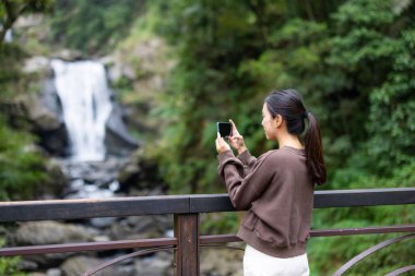 Kadın cep telefonuyla şelaleyle fotoğraf çekiyor.