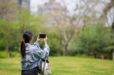 Kadın parkta fotoğraf çekmek için cep telefonu kullanıyor.