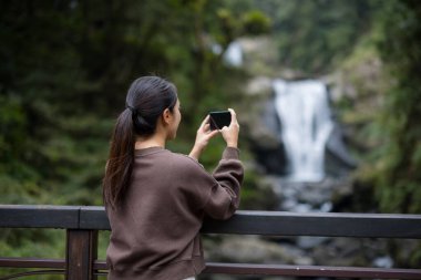 Kadın ormanda şelaleyle fotoğraf çekmek için cep telefonu kullanıyor.