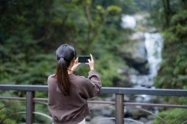 Kadın şelalede fotoğraf çekmek için cep telefonu kullanıyor.
