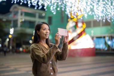 Kadın cep telefonuyla şehrin Noel dekorasyon geçmişiyle fotoğraf çekiyor.