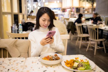 Kadın lokantada yemek yerken cep telefonu kullanıyor. 