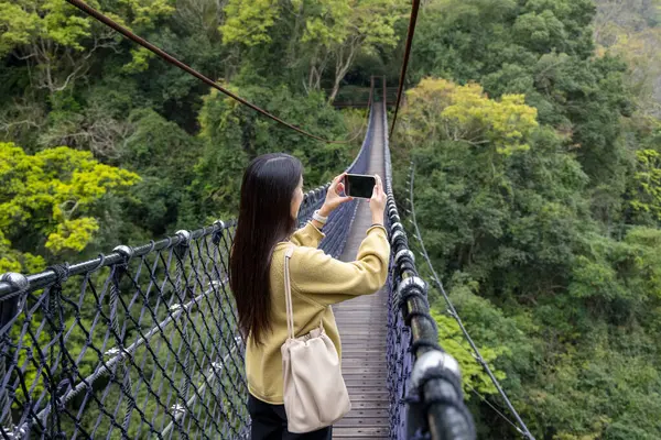Woman walk along the suspension bridge and use mobile phone to take photo in the forest