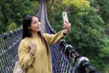 Kadın asma köprü boyunca yürür ve ormanda fotoğraf çekmek için cep telefonu kullanır.