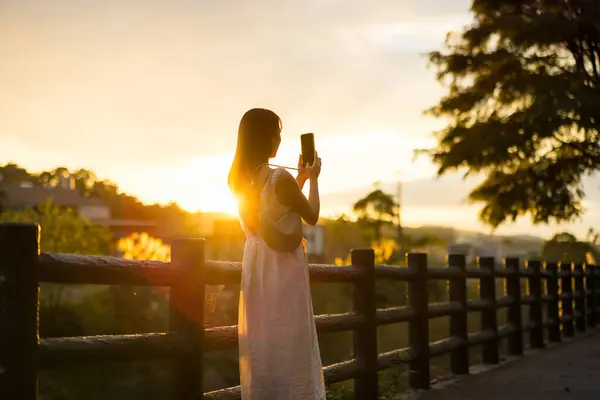 女性は田舎の日没の下で写真を撮るために携帯電話を使用します ストック写真
