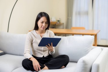 Asyalı kadın dijital tablet bilgisayar kullanıyor ve evdeki kanepeye oturuyor.