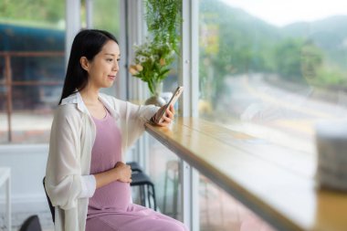 Hamile kadın kahve dükkanında oturuyor ve cep telefonu kullanıyor.