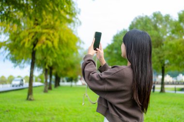 Kadın parkta fotoğraf çekmek için cep telefonu kullanıyor.