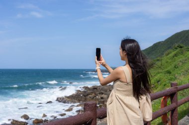 Hamile kadın deniz kenarında fotoğraf çekmek için cep telefonu kullanıyor.