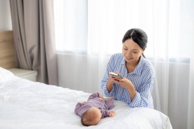 Asyalı anne bebeğinin fotoğrafını çekmek için cep telefonu kullanıyor.