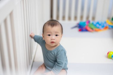 Beyaz beşiğin içinde oturan meraklı bebek etrafa bakıyor.