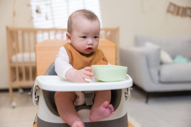 Bebek sandalyeye oturmuş, kaseyi tutuyor.