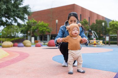 Anne, bebeğin ayı şapkasıyla parkta yürümesine yardım ediyor.