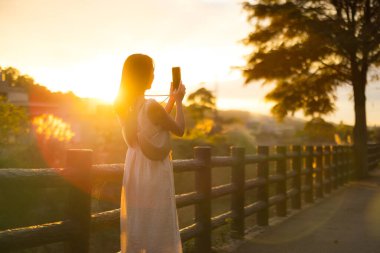 Dışarıda gün batımı fotoğrafı çekmek için cep telefonu kullanan bir kadın.