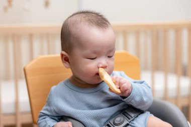 Küçük Asyalı bebek sandalyede pirinç krakerinin tadını çıkarıyor.