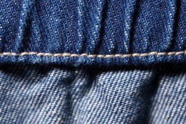 Macro image of blue denim fabric showing detailed stitching, seam, and textured weave pattern.