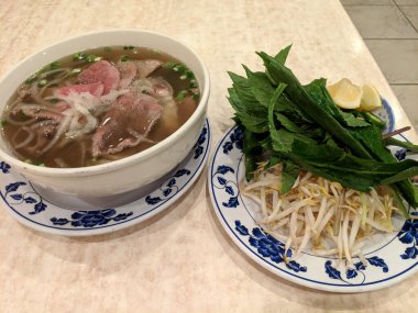 Biftek dilimleri, soğanlar, taze otlar, fasulye filizleri, limon dilimleri ve soğanlı Pho 'nun (Vietnam Pirinç Erişte Çorbası) yakın çekimi..