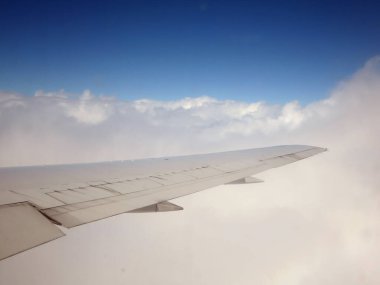 Gökyüzünde yüksek uçan ticari bir jet uçağının penceresinden görülen tüylü beyaz bulutların hava görüntüsü. Uçağın kanadı görüntüde görülebilir..