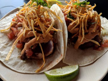 Un tortilla, lahana, limon, mısır şeridi, pico de gallo ve imzalı aioli ile yapılmış lezzetli ve taze bir balık tacosu. Tacolar yerel olarak yakılmış, az pişmiş ahi ton balığıyla dolu, beyaz tabakta servis ediliyor.