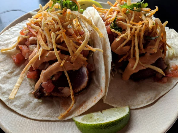 A delicious and fresh dish of fish tacos, made with flour tortillas, cabbage, lime, corn strips, pico de gallo, and signature aioli. The tacos are filled with locally caught seared rare ahi tuna, served on a white plate