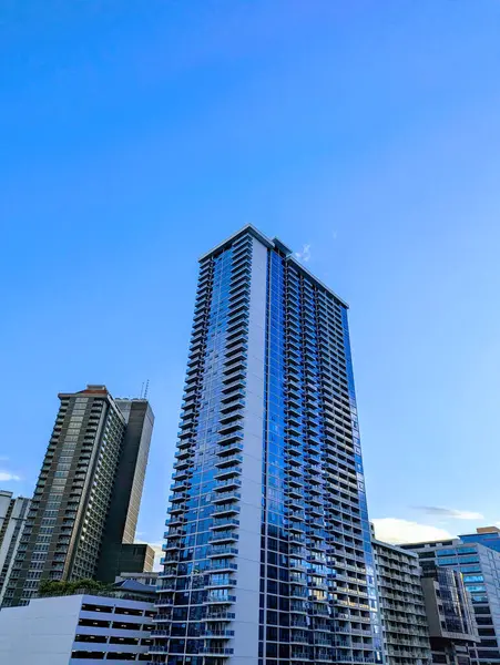 Honolulu - 17 Temmuz 2023: Oahu, Hawaii 'deki Ala Moana bölgesindeki modern yüksek katlı bir apartmanın fotoğrafı. Bina uzun ve dikdörtgen ve mavi cam cepheli. Fotoğraf, şehir ve doğal manzara arasındaki zıtlığı gösteriyor. 