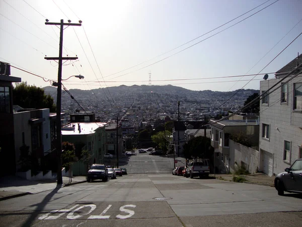 San Francisco - 26 Mart 2010: San Francisco, Kaliforniya 'da bir caddenin parlak ve neşeli bir fotoğrafı. Sokak aşağı doğru eğimli ve arka planda şehrin ufuk çizgisinin panoramik bir görüntüsü var. Cadde beyaz evler ve araba parklarıyla dolu.
